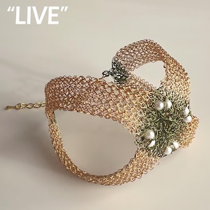 May include: A gold-toned metal mask with a chainmail design. The mask features a decorative section with green accents and white pearl embellishments. The word "LIVE" is printed in white at the top. The mask has a chain closure.