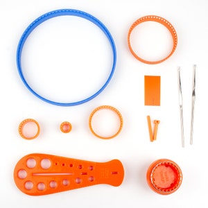 May include: A set of orange and blue embroidery hoops, a ruler with multiple sized circles, two crochet hooks, and a small orange rectangle. The hoops are made of plastic and have a textured surface. The ruler is made of plastic and has a smooth surface. The crochet hooks are made of metal and have a smooth surface. The rectangle is made of plastic and has a smooth surface.