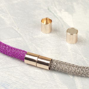 Op de afbeelding: Een close-up van een magnetische sluiting voor een ketting. De sluiting is gemaakt van twee goudkleurige metalen delen die in elkaar passen. De ketting is gemaakt van een zilveren en paarse mesh-stof.