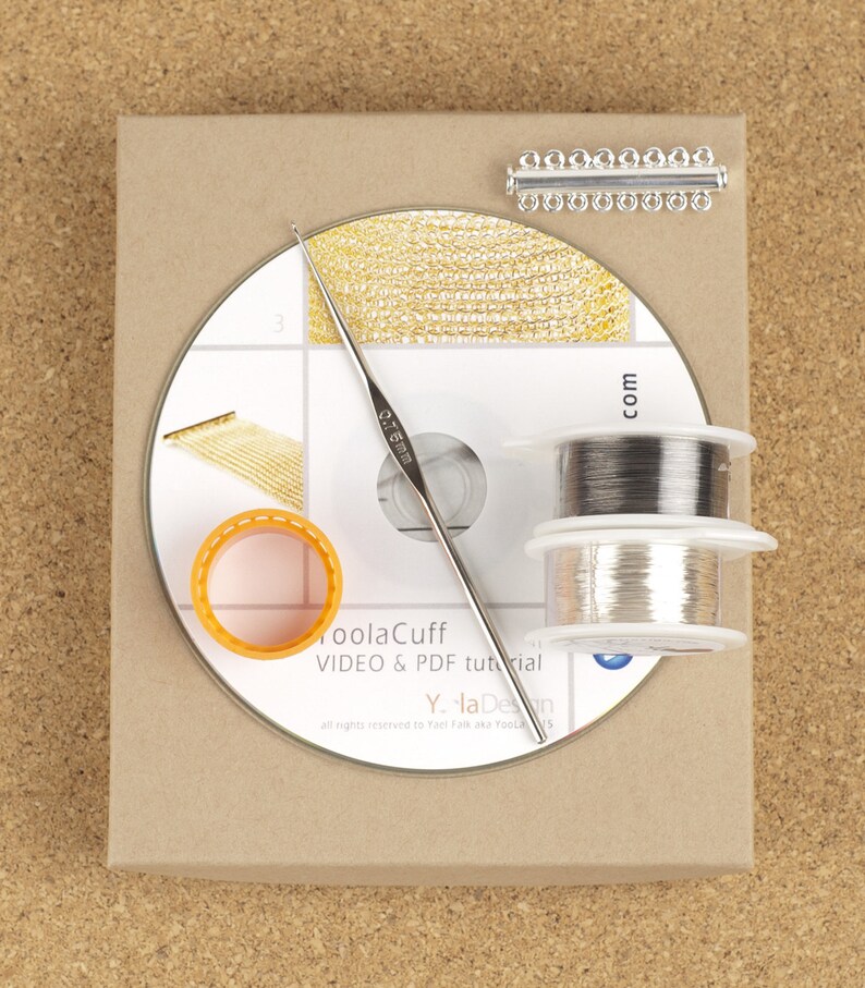 Puede incluir: Una caja marr&oacute;n con un CD blanco en su interior. El CD tiene una cadena dorada, un gancho plateado y dos carretes de alambre plateado en la parte superior. La caja tiene el texto "ToolaCuff VIDEO & PDF tutorial Y. la Design all rights reserved to Yael Falk aka Yoola 15 com" impreso en ella.