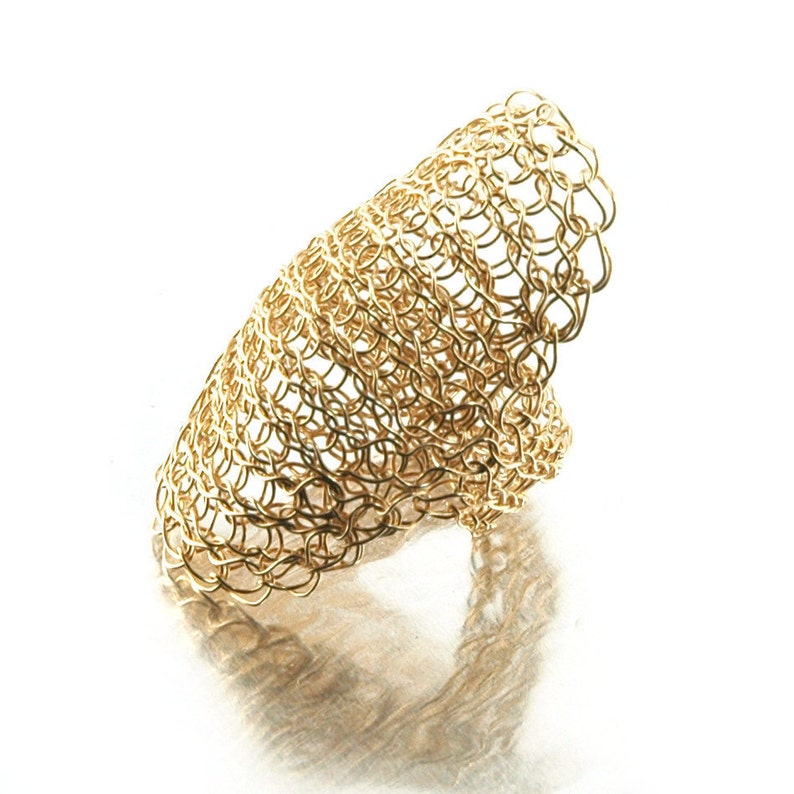 May include: A gold-colored metal ring with a woven design. The ring is shaped like a curved shell.