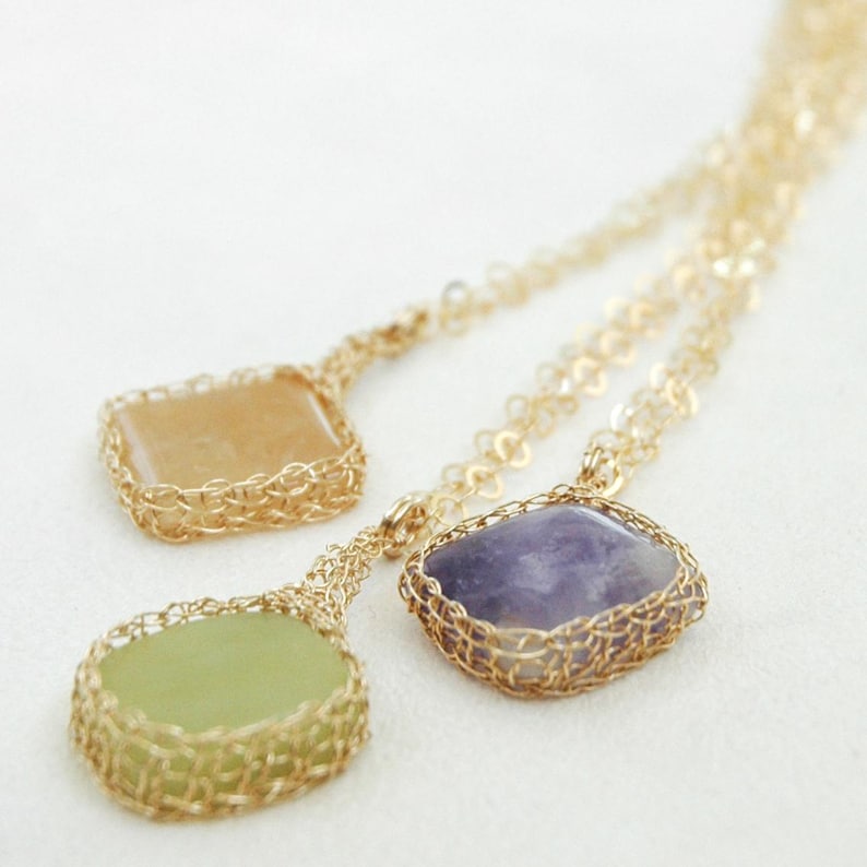 May include: Three gold necklaces with different colored gemstone pendants. The pendants are a square of light brown, a square of purple, and a round green stone. Each stone is encased in a gold wire mesh.