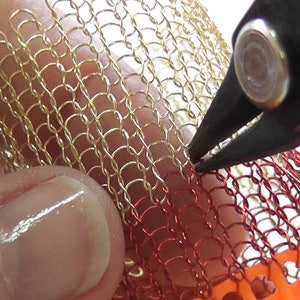 Puede incluir: Primer plano de un par de alicates sujetando una malla de alambre dorada y roja. La malla se est&aacute; utilizando para crear una pieza de joyer&iacute;a.