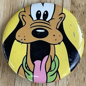Op de afbeelding: Een ronde knoop met Pluto, een Disney-tekenfilmhond, op een gele achtergrond. Pluto's gezicht is bruin met zwarte oren, een zwarte neus en een roze tong. De knoop heeft een groene kraag en het Disney-copyrightsymbool.