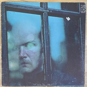 Puede incluir: Una portada de álbum de vinilo vintage con la cara de un hombre detrás de un cristal de ventana. La portada del álbum es azul y negra con el texto "TOM PAXTON 6" en blanco.