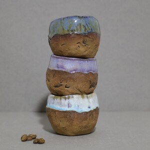 Puede incluir: Una pila vertical de tres tazas de cerámica pequeñas, redondas y hechas a mano, con una base de arcilla marrón rugosa sin esmaltar y un borde esmaltado liso y brillante en tonos azul, morado y blanco, junto a unos granos de café esparcidos.