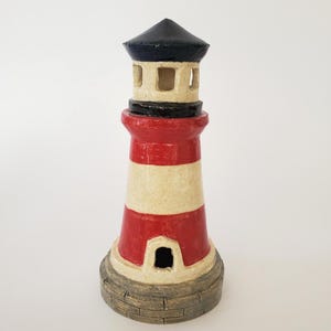 Puede incluir: Un faro de cerámica con una parte superior negra, rayas rojas y crema, y una base texturizada. El faro tiene una pequeña abertura cerca de la base y cortes cuadrados cerca de la parte superior. Un objeto náutico decorativo.