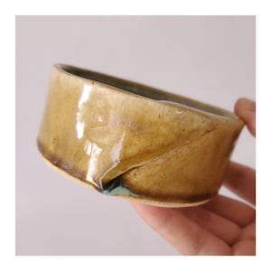 Puede incluir: Un cuenco de cerámica hecho a mano con un diseño plegado único. El cuenco presenta una paleta de colores cálidos y terrosos con tonos de beige y marrón, y un toque de verde azulado en el interior. El cuenco está sostenido por una mano.