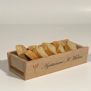 Puede incluir: Una cesta de pan de madera con una mancha marrón y el texto "Agriturismo Il Mulino" grabado en la parte delantera. La cesta está llena de rebanadas de pan.