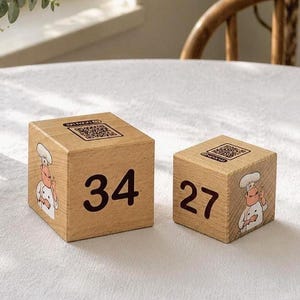 Puede incluir: Dos bloques de madera con los números 34 y 27 impresos en negro. Cada bloque presenta un dibujo de un chef y un código QR. Los bloques son de color marrón claro y están sobre una superficie blanca.