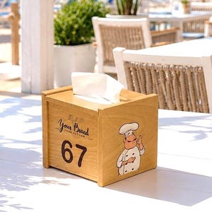 Può includere: Scatola di fazzoletti di legno con la scritta "Your Brand" e il numero "67". Un cuoco dei cartoni animati è sul lato. La scatola è su un tavolo bianco, con una scena di spiaggia sullo sfondo.