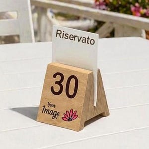 Può includere: Portanumero da tavolo in legno con il numero 30 in marrone. Il supporto ha una fessura per un cartello riservato. La base ha il testo "Your Image" e un disegno floreale rosa. Il supporto è su un tavolo bianco.