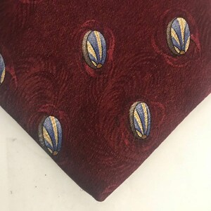 Peut inclure: Gros plan sur une cravate bordeaux avec un motif répété de formes ovales bleues et dorées. La cravate a une surface texturée et un design classique, adapté aux tenues formelles ou professionnelles.