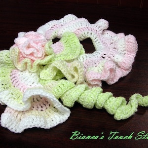 May include: Handmade crochet floral embellishments in pastel colors. The design features a pink flower, white and pink ruffles, and a green spiral. The text "Bianca's Touch Studio" is visible in the image.