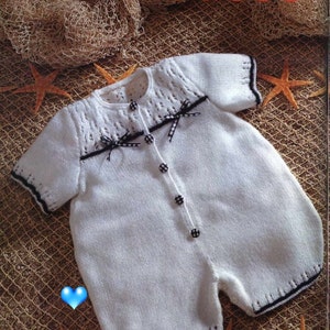 May include: White knitted baby romper with black trim and black and white checked buttons. The romper has a delicate lace pattern on the neckline and shoulders. The romper is photographed against a brown net background.