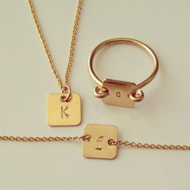 May include: A set of three gold-toned jewelry pieces: a necklace with a square pendant engraved with the letter "K", a bracelet with a square pendant engraved with the letter "F", and a ring with a square pendant engraved with the letter "C".
