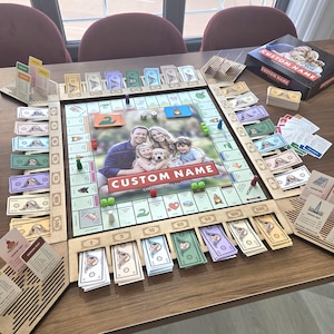 May include: A custom board game set featuring a central square game board with a family photograph and the text CUSTOM NAME, surrounded by wooden card holders, stacks of play money, game pieces, dice, and a box labeled CUSTOM NAME.