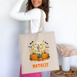 May include: Beige Halloween tote bag featuring a skeleton dancing in front of a full moon, pumpkins, and falling leaves. The name "NATALIE" is printed in orange. Black bats are flying around the moon. The bag has a simple design.