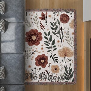 Può includere: Una coperta in tessuto bianco con un motivo floreale con fiori e foglie rossi, marroni e beige. La coperta ha frange su tutti i lati e il testo "The Robinsons" è stampato al centro in basso.