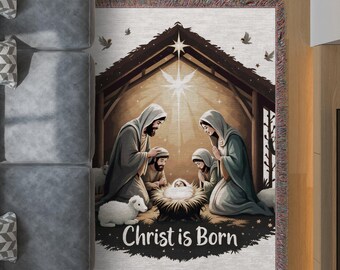 Op de afbeelding: Een geweven sprei met een kerststalscène met de tekst "Christ is Born". De afbeelding toont Maria, Jozef en het kindje Jezus in een kribbe, met een heldere ster erboven. De sprei heeft een gefranjerde rand en een neutraal kleurenpalet.