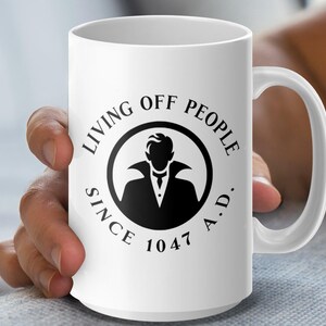 Könnte beinhalten: Weiße Keramik-Kaffeetasse mit einer schwarzen Grafik eines Vampirs mit dem Text "Living off people since 1047 A.D."