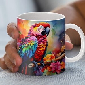 Könnte beinhalten: Eine weiße Keramiktasse mit einem bunten Papageien-Design. Der Papagei sitzt auf einem Ast mit rosa Blüten. Der Hintergrund ist ein Aquarell-Stil-Gemälde mit Farbspritzern.