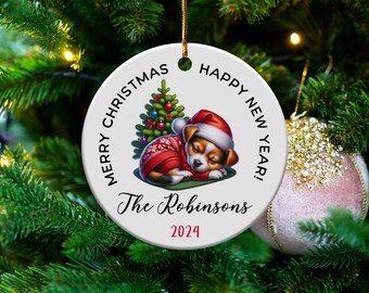 Könnte beinhalten: Runder weißer Weihnachtsbaumschmuck mit einem schlafenden Welpen, der eine rote Weihnachtsmütze und einen rot-weißen Pullover trägt. Der Anhänger hat den Text "Frohe Weihnachten Frohes Neues Jahr! Die Robinsons 2024" aufgedruckt.