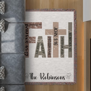 Op de afbeelding: Een witte geweven deken met het woord "FAITH" in grote letters, waarbij elke letter een ander Bijbelvers voorstelt. De verzen zijn: Hebreeën 11:1, Mattheüs 21:22, Johannes 3:16, Spreuken 3:5-6, Jakobus 2:17, Psalm 37:5, Efeziërs 2:8, Romeinen 10:17. De deken heeft ook de tekst "The Robinsons" onderaan.
