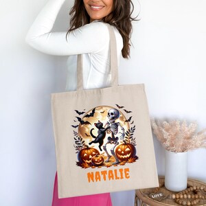 Puede incluir: Bolso tote de lona beige con un diseño de Halloween. El diseño incluye un esqueleto, un gato negro, calabazas, murciélagos y una luna llena. El nombre "NATALIE" está impreso en naranja. El bolso tiene dos asas.