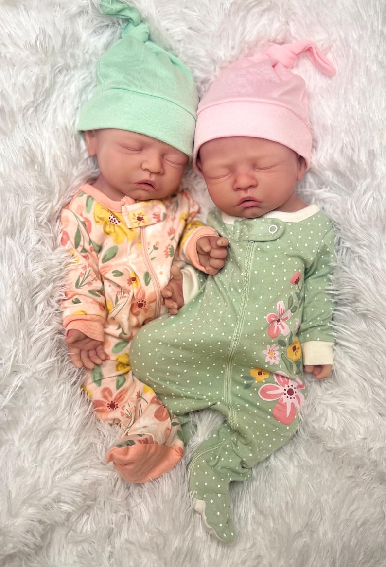 May include: Two realistic baby dolls wearing matching knotted hats. One doll is in a floral print jumpsuit, the other in a green polka dot jumpsuit with floral accents. Both dolls are lying on a white, fluffy surface.