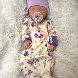 K&ouml;nnte beinhalten: Ein Baby mit einer hellvioletten M&uuml;tze mit Ohren und einem cremefarbenen Strampler mit lila Eulen- und Baummustern. Das Baby hat einen Schnuller im Mund und liegt auf einer flauschigen wei&szlig;en Oberfl&auml;che.