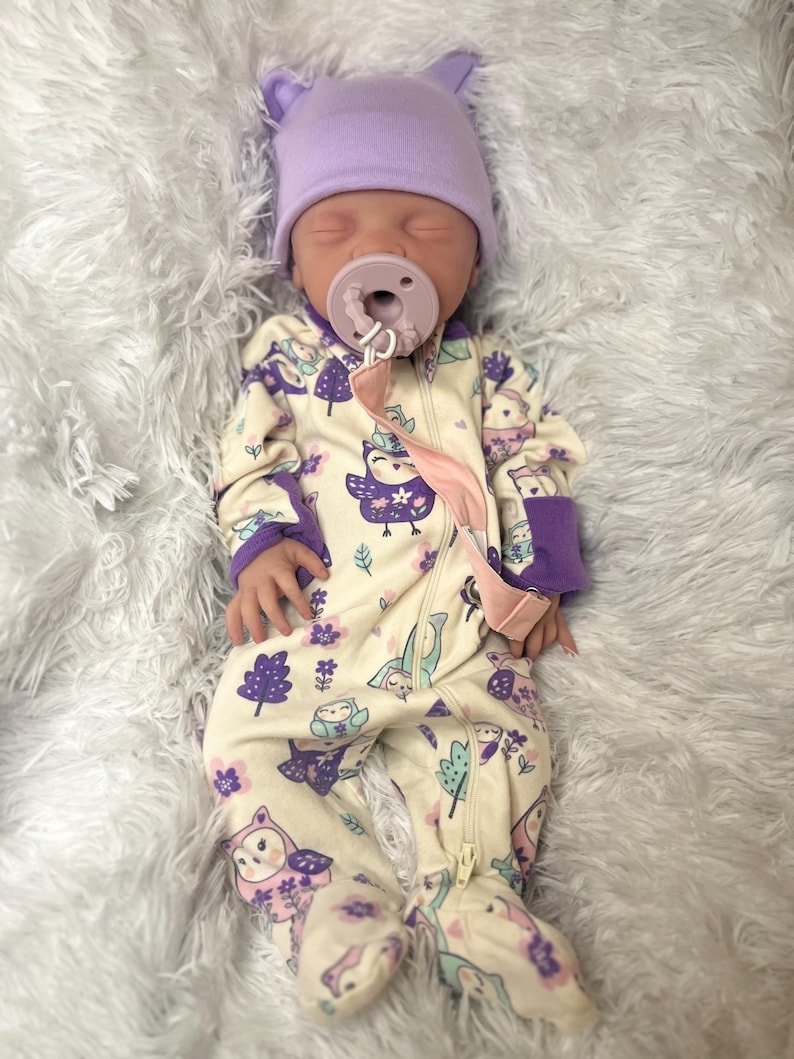 K&ouml;nnte beinhalten: Ein Baby mit einer hellvioletten M&uuml;tze mit Ohren und einem cremefarbenen Strampler mit lila Eulen- und Baummustern. Das Baby hat einen Schnuller im Mund und liegt auf einer flauschigen wei&szlig;en Oberfl&auml;che.
