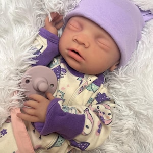 K&ouml;nnte beinhalten: Eine realistische Babypuppe mit einer lavendelfarbenen Strickm&uuml;tze und einem gemusterten Strampler mit lila B&uuml;ndchen. Die Puppe hat einen Schnuller und liegt auf einer flauschigen wei&szlig;en Oberfl&auml;che.