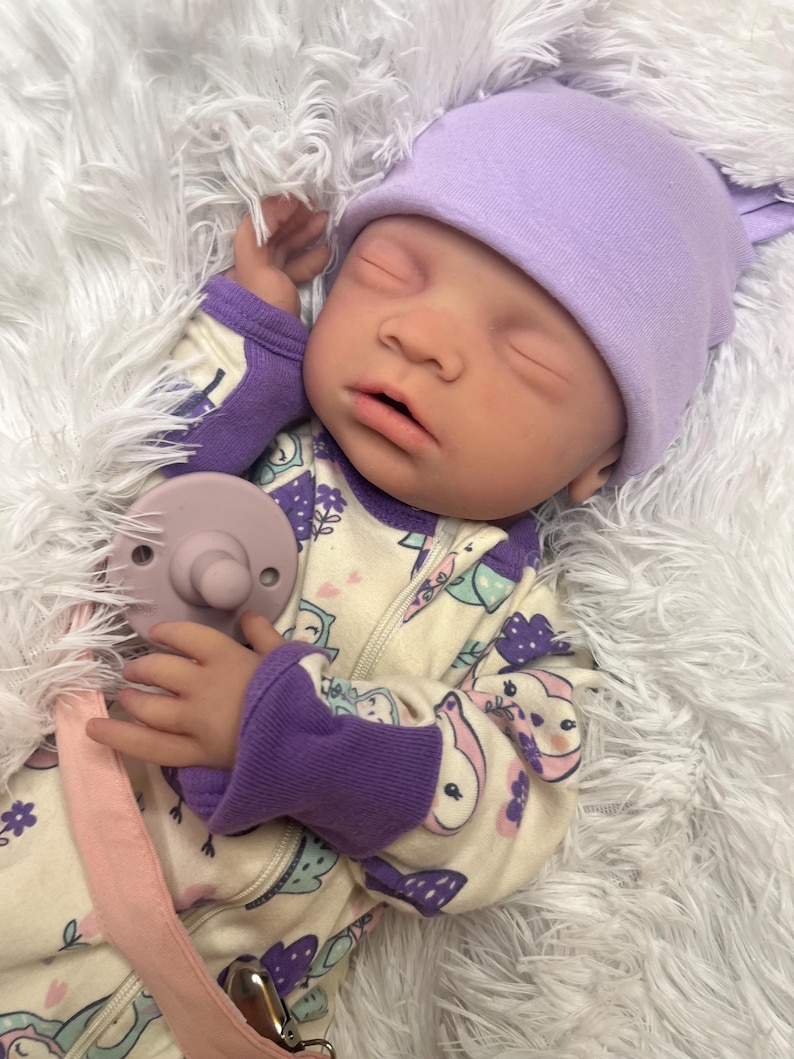 K&ouml;nnte beinhalten: Eine realistische Babypuppe mit einer lavendelfarbenen Strickm&uuml;tze und einem gemusterten Strampler mit lila B&uuml;ndchen. Die Puppe hat einen Schnuller und liegt auf einer flauschigen wei&szlig;en Oberfl&auml;che.