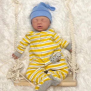 K&ouml;nnte beinhalten: Ein Baby mit blauer M&uuml;tze und einem gelb-wei&szlig; gestreiften Strampler mit Faultiermotiv, das auf einer Holzschaukel liegt. Das Baby liegt auf einer flauschigen wei&szlig;en Oberfl&auml;che.