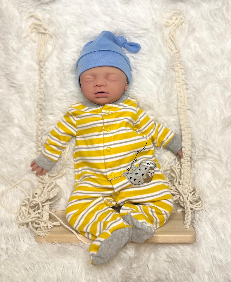 K&ouml;nnte beinhalten: Ein Baby mit blauer M&uuml;tze und einem gelb-wei&szlig; gestreiften Strampler mit Faultiermotiv, das auf einer Holzschaukel liegt. Das Baby liegt auf einer flauschigen wei&szlig;en Oberfl&auml;che.
