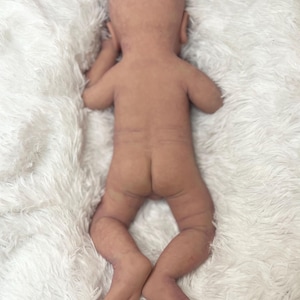 K&ouml;nnte beinhalten: Eine realistische, nackte Babypuppe liegt auf einer flauschigen, wei&szlig;en Decke. Die Puppe liegt auf dem Bauch, Arme und Beine sind leicht angewinkelt. Die Haut der Puppe hat einen realistischen Ton und eine realistische Textur.