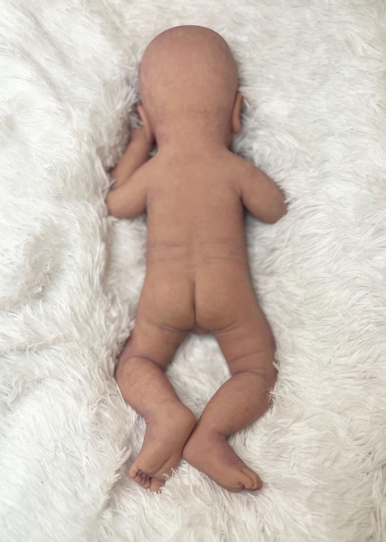 K&ouml;nnte beinhalten: Eine realistische, nackte Babypuppe liegt auf einer flauschigen, wei&szlig;en Decke. Die Puppe liegt auf dem Bauch, Arme und Beine sind leicht angewinkelt. Die Haut der Puppe hat einen realistischen Ton und eine realistische Textur.