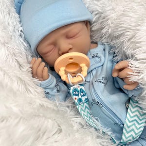May include: A realistic baby doll wearing a light blue hat and onesie with a dinosaur pattern. The doll has a pacifier and is lying on a fluffy white surface. The pacifier clip has a blue and white chevron pattern.
