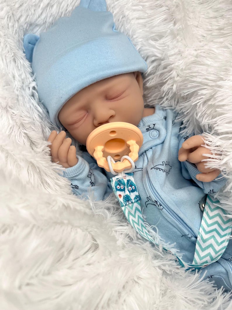 May include: A realistic baby doll wearing a light blue hat and onesie with a dinosaur pattern. The doll has a pacifier and is lying on a fluffy white surface. The pacifier clip has a blue and white chevron pattern.