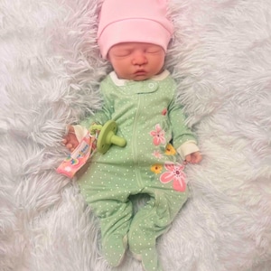 May include: A newborn baby wearing a light green onesie with white polka dots and floral accents, and a pink knotted cap. The baby is lying on a fluffy white blanket, with a pacifier in its mouth.