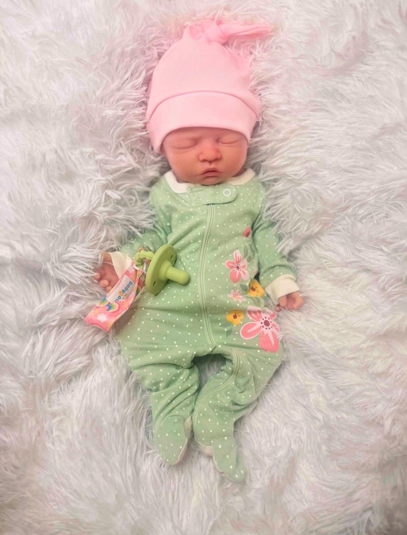 May include: A newborn baby wearing a light green onesie with white polka dots and floral accents, and a pink knotted cap. The baby is lying on a fluffy white blanket, with a pacifier in its mouth.