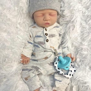 May include: A lifelike baby doll wearing a gray bear-eared hat and a light gray onesie with a cloud and airplane pattern. The doll has a blue pacifier attached to a black and white polka dot strap.