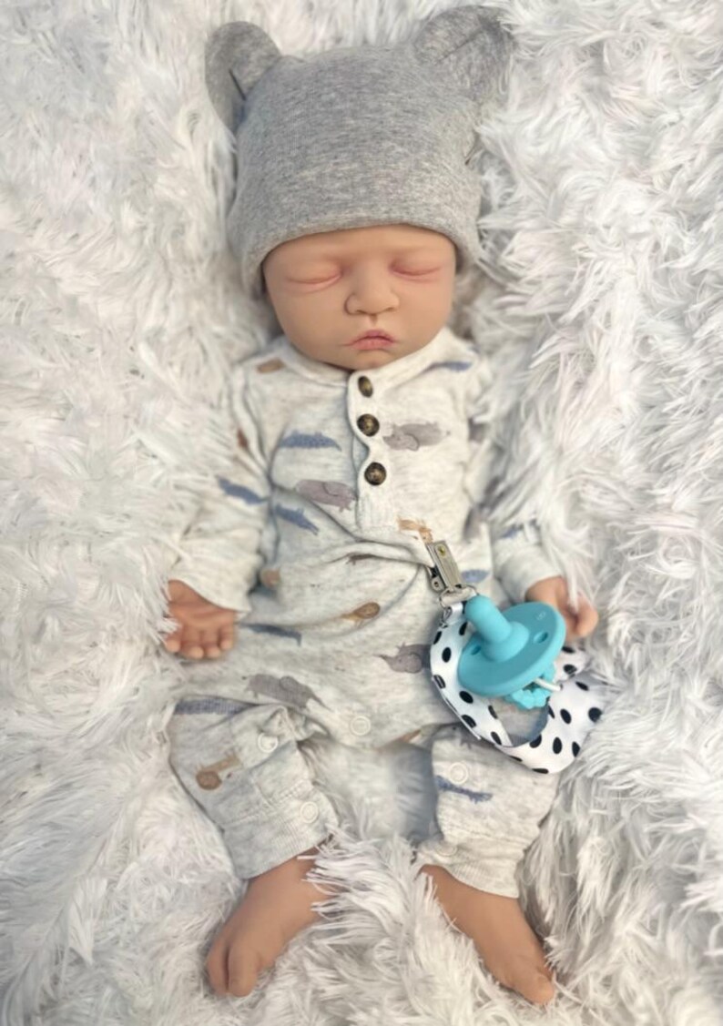 May include: A lifelike baby doll wearing a gray bear-eared hat and a light gray onesie with a cloud and airplane pattern. The doll has a blue pacifier attached to a black and white polka dot strap.