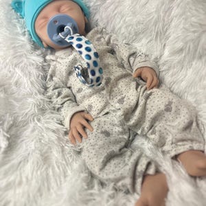 K&ouml;nnte beinhalten: Eine Babypuppe mit einer hellblauen M&uuml;tze mit B&auml;renohren, einem grauen Strampler mit einem Muster aus kleinen grauen Formen und einem blauen Schnuller. Der Schnuller hat einen blau-wei&szlig; gepunkteten Riemen.