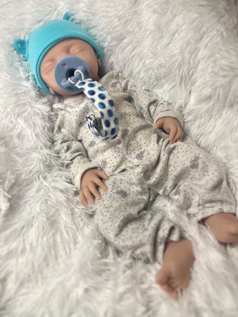K&ouml;nnte beinhalten: Eine Babypuppe mit einer hellblauen M&uuml;tze mit B&auml;renohren, einem grauen Strampler mit einem Muster aus kleinen grauen Formen und einem blauen Schnuller. Der Schnuller hat einen blau-wei&szlig; gepunkteten Riemen.