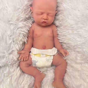 May include: A lifelike baby doll with a bald head and closed eyes, lying on a fluffy white surface. The doll has a realistic skin tone and is wearing a diaper with an elephant print. The baby's arms and legs are slightly bent.