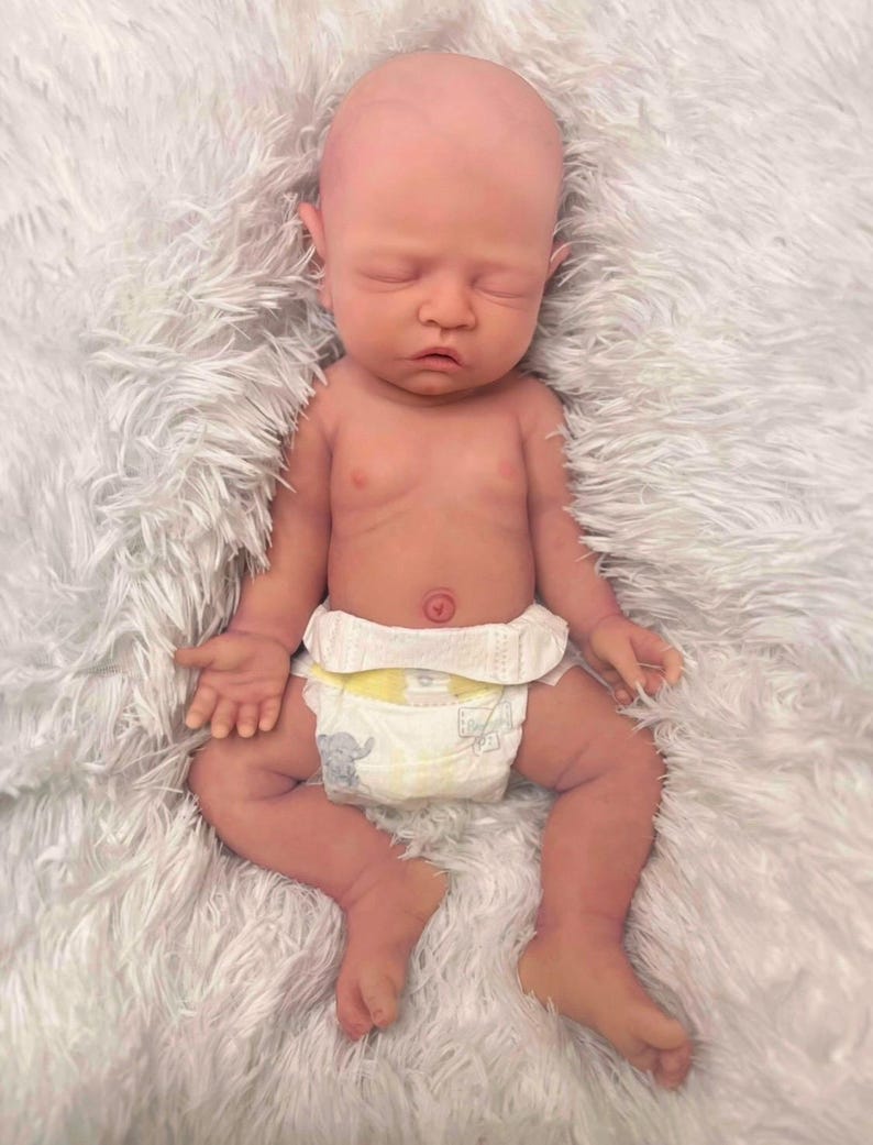 May include: A lifelike baby doll with a bald head and closed eyes, lying on a fluffy white surface. The doll has a realistic skin tone and is wearing a diaper with an elephant print. The baby's arms and legs are slightly bent.