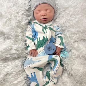 K&ouml;nnte beinhalten: Ein Baby mit einem grauen Hut mit kleinen Ohren und einem wei&szlig;en Strampler mit blauem und gr&uuml;nem Dinosaurier-Muster. Das Baby h&auml;lt auch einen blauen Schnuller. Das Baby liegt auf einer flauschigen wei&szlig;en Oberfl&auml;che.