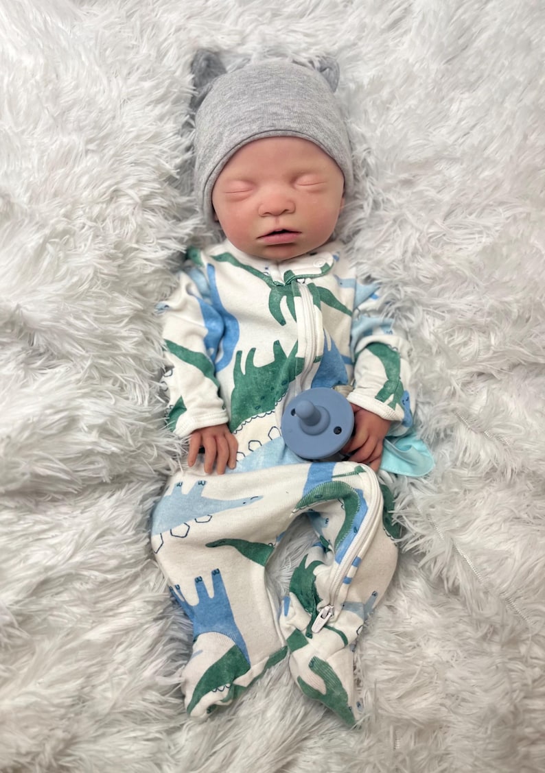 K&ouml;nnte beinhalten: Ein Baby mit einem grauen Hut mit kleinen Ohren und einem wei&szlig;en Strampler mit blauem und gr&uuml;nem Dinosaurier-Muster. Das Baby h&auml;lt auch einen blauen Schnuller. Das Baby liegt auf einer flauschigen wei&szlig;en Oberfl&auml;che.